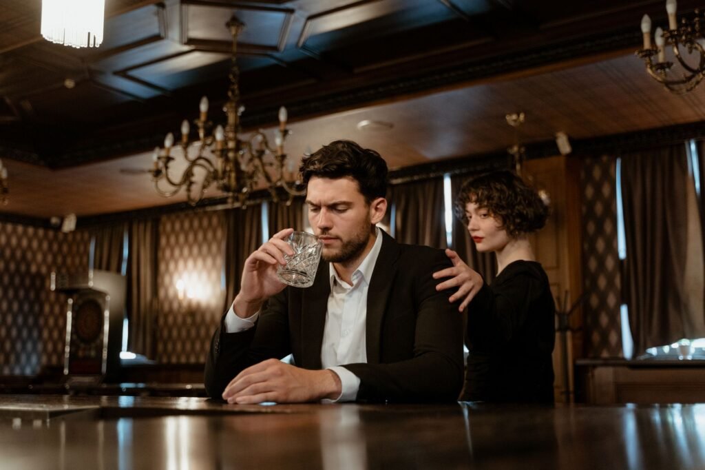 An elegant bar scene with a thoughtful man holding a glass and a woman comforting him.