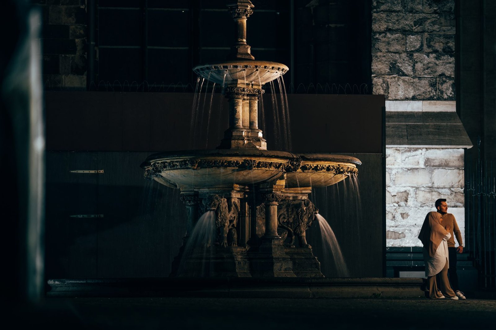 A couple embraces by a beautifully lit ornate fountain at night, capturing a classic romantic scene.