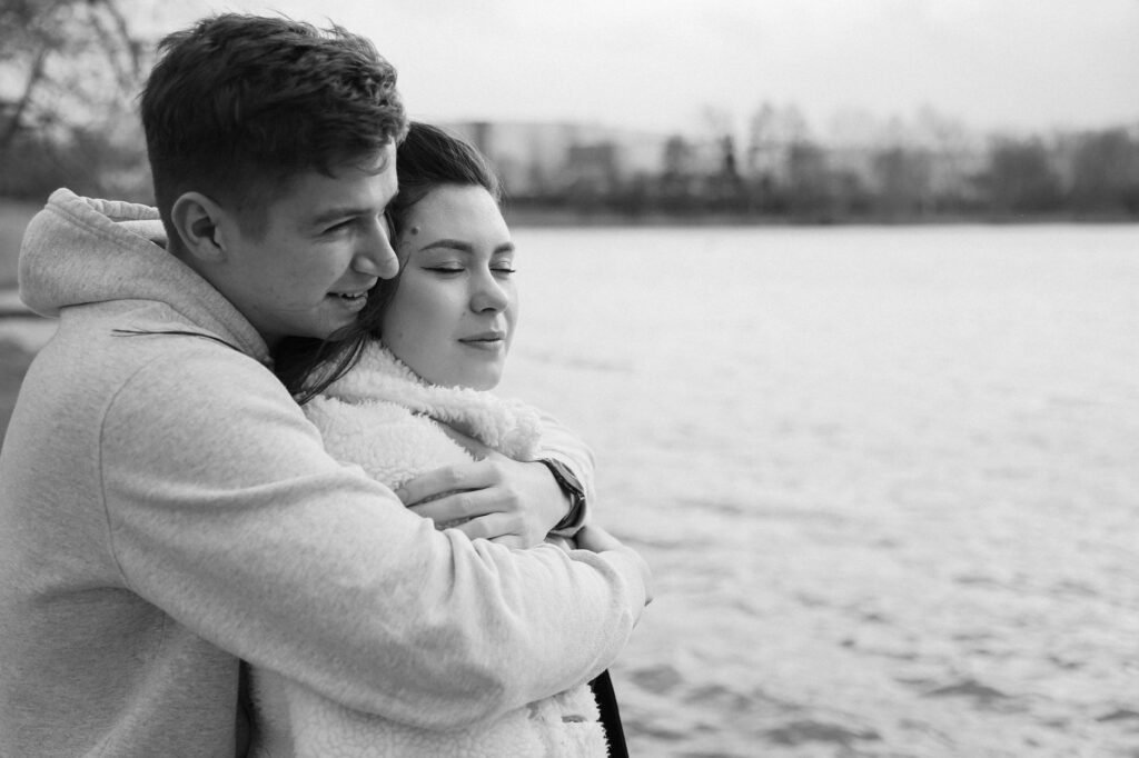 A romantic black and white photograph of a couple embracing by the water, evoking warmth and togetherness.