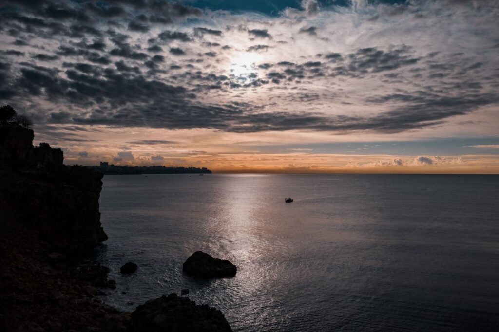 Dramatic sunset over the calm ocean framed by a rocky cliff silhouette and scattered clouds.