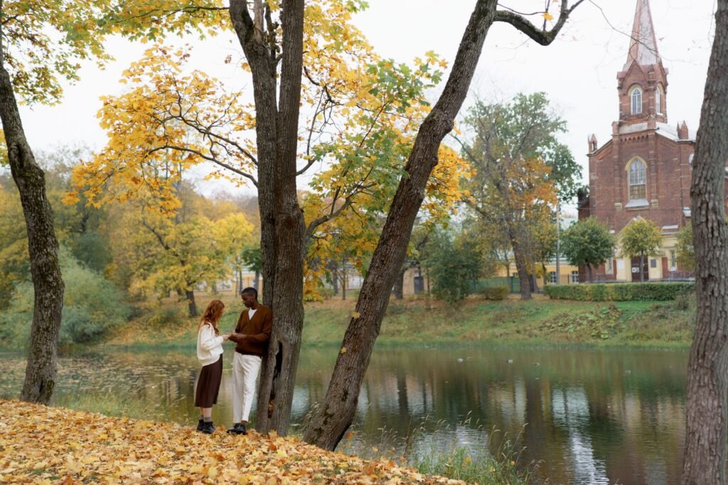 A multiracial couple enjoys a walk by a lake surrounded by fall colors and historic architecture.