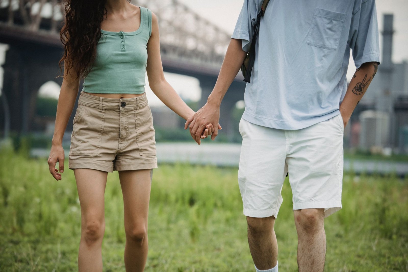 Unrecognizable couple in casual wear holding hands and walking on grassy lawn on street with bridge on blurred background in
