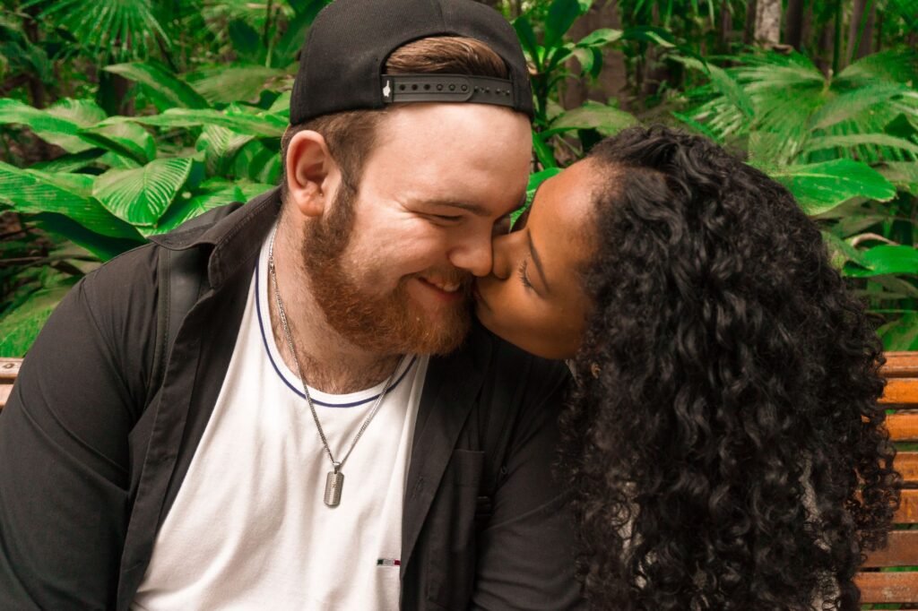 A loving couple kisses tenderly while seated outdoors in lush greenery, showcasing affection and intimacy.