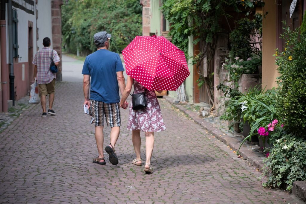 Couple walking under a red polka-dot umbrella on a cobblestone street in Kaysersberg, France.