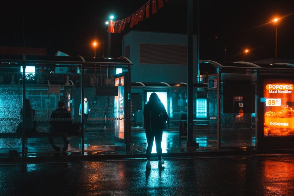 Silhouetted figures wait at an illuminated bus stop on a rainy night, capturing urban nightlife's mood.