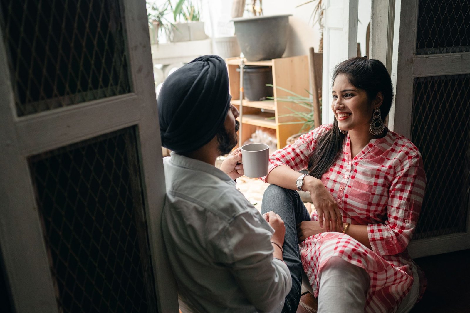 Cheerful Indian couple of smiling lady listening to boyfriend telling amusing story and drinking coffee on floor at threshold