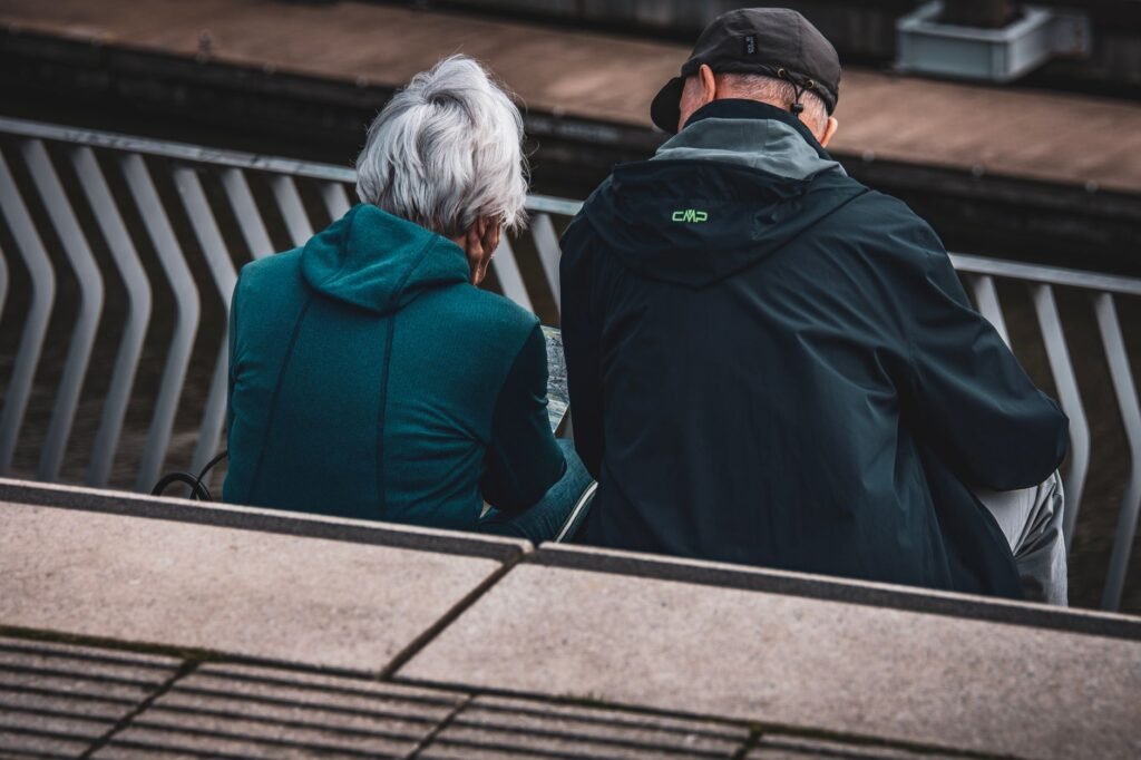 Elderly couple in jackets sitting on steps overlooking an urban setting, captured candidly from behind.
