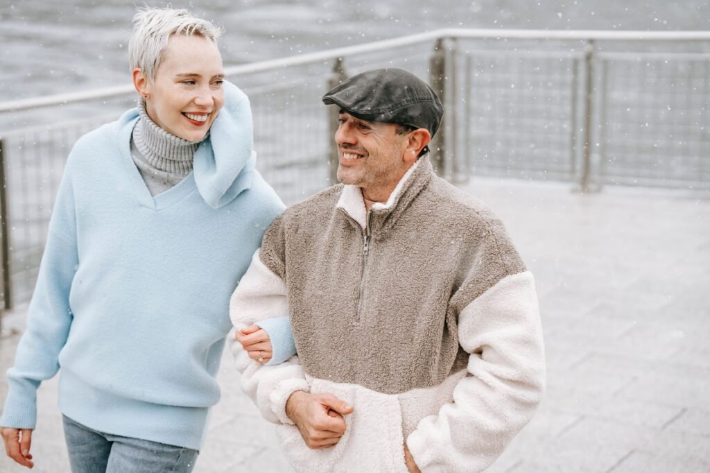 Cheerful couple in warm clothes talking on pavement against river in city in cold weather