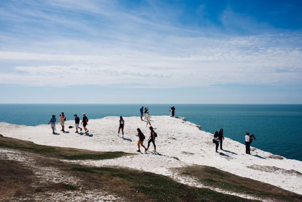 A group of people walk along dramatic white cliffs overlooking the ocean under a blue sky.