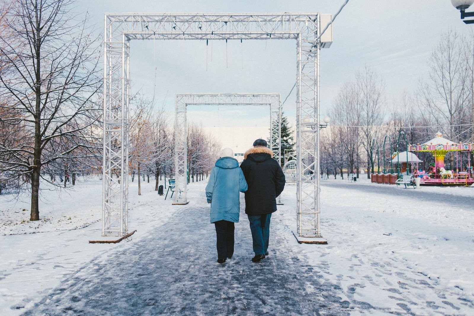 Back view of anonymous couple strolling on snowy walkway with rectangular shaped arches in amusement park in town