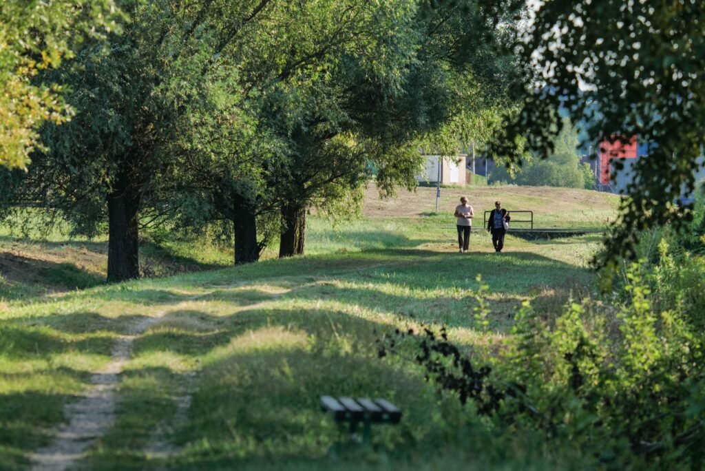 Two people walking on a green rural path in Garešnica, surrounded by trees on a sunny day.