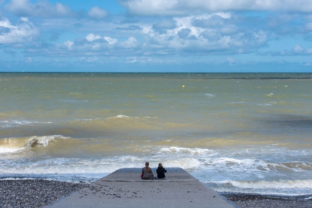 Two individuals sit on a concrete jetty overlooking a calm sea under a partly cloudy sky.