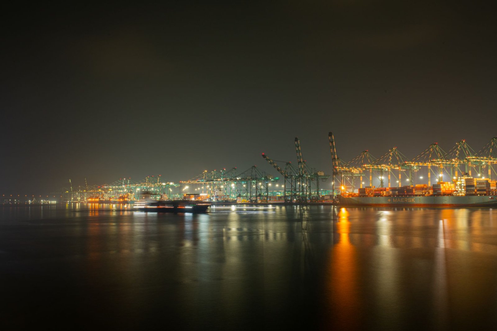 A vibrant night view of a busy cargo port with cranes and ships reflecting on the water under city lights.