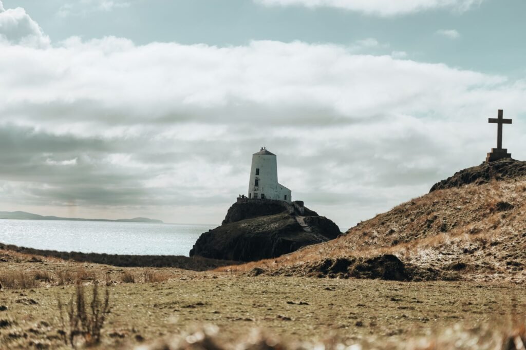 Scenic view of Twr Mawr Lighthouse and a cross on Ynys Llanddwyn Island with sea and cloudy sky.