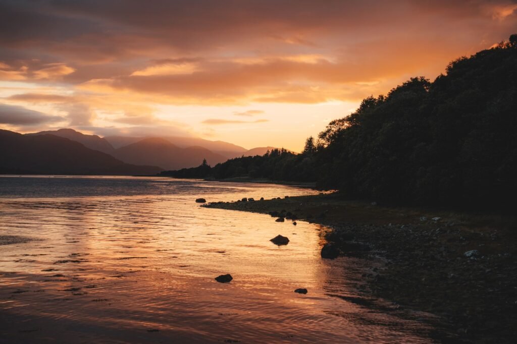 Captivating sunset over a serene loch with silhouettes of mountains and trees in Scotland.