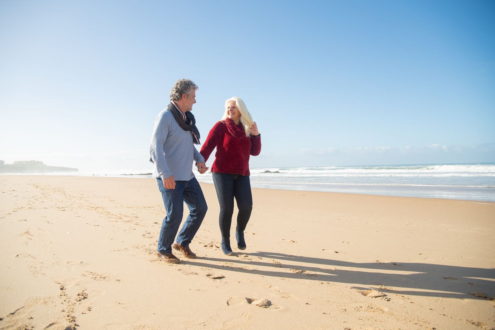 A happy senior couple holds hands while walking along a sunny beach in Portugal, enjoying a romantic moment.