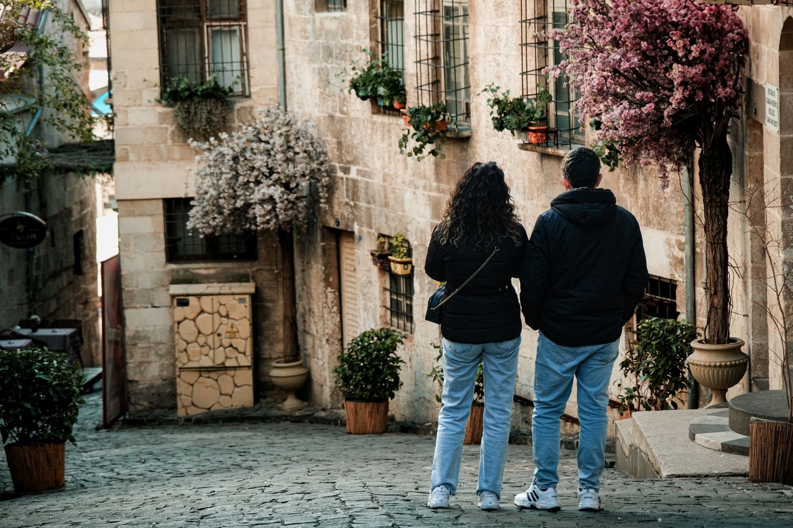 A couple casually walks down a charming, flower-adorned cobblestone street in a historic area.