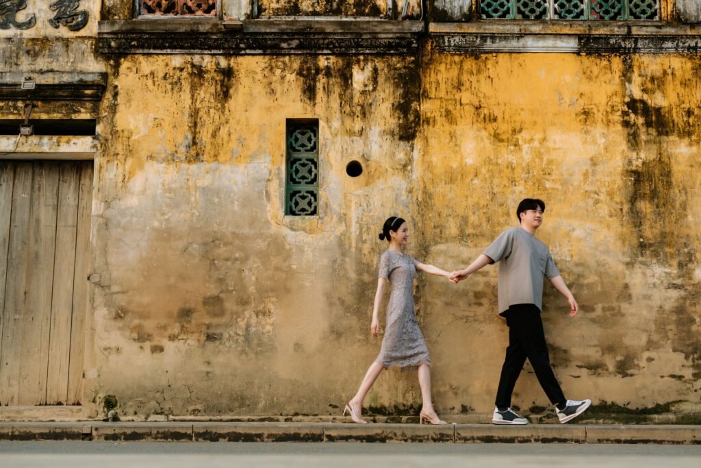 A couple holding hands, walking along a rustic yellow wall, creating a romantic street scene.