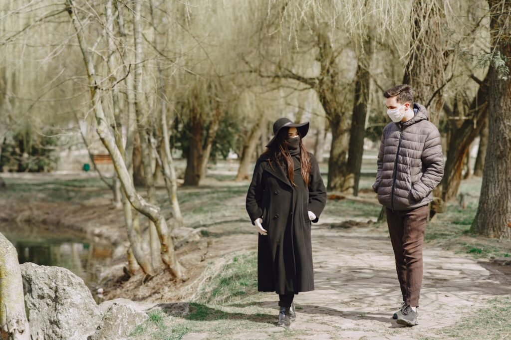 Full body young couple in outerwear and protective masks walking along path in empty park during COVID 19 outbreak and chatti