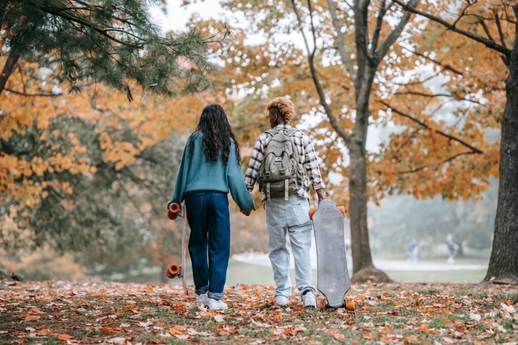 A young couple holding hands and carrying skateboards in a vibrant autumn park setting.