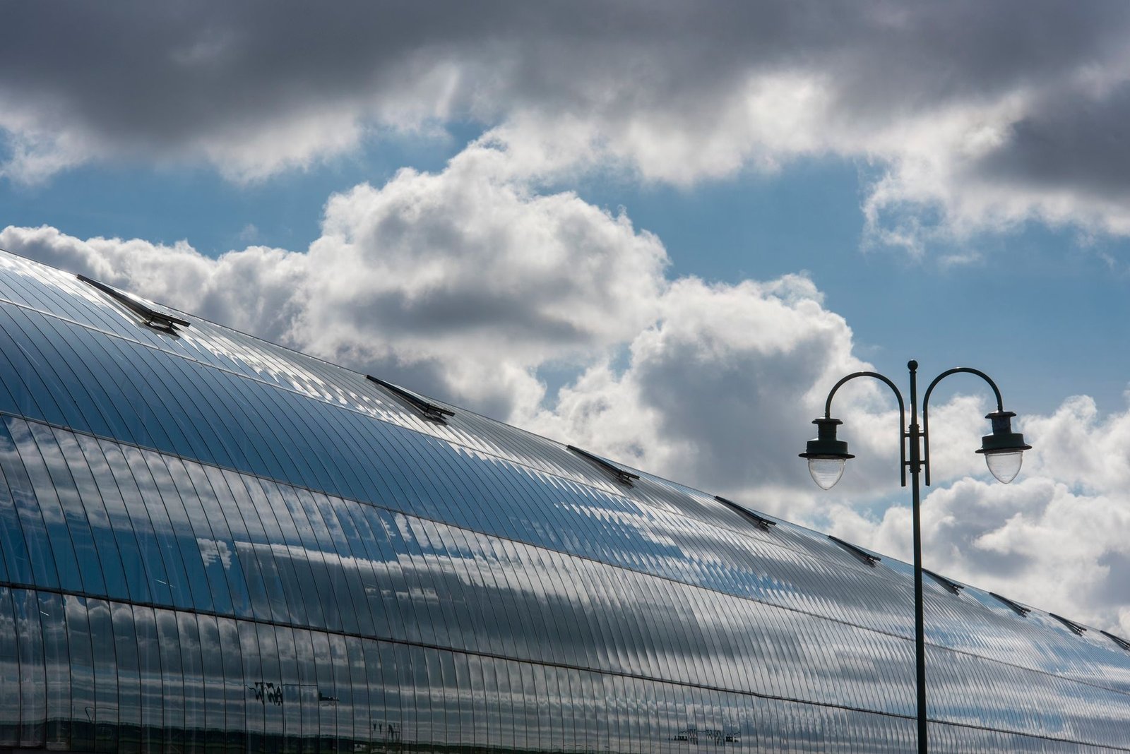 Reflective curved building facade under cloudy sky with streetlamp. Modern architecture and weather contrast.