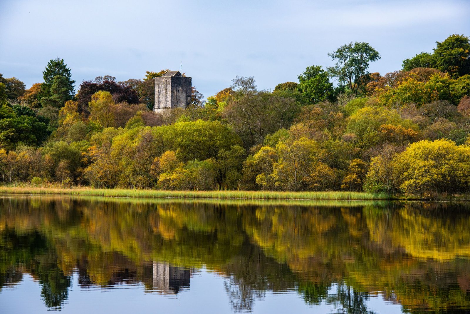Tranquil view of Mugdock Loch reflecting autumn foliage and a historical tower in Scotland.