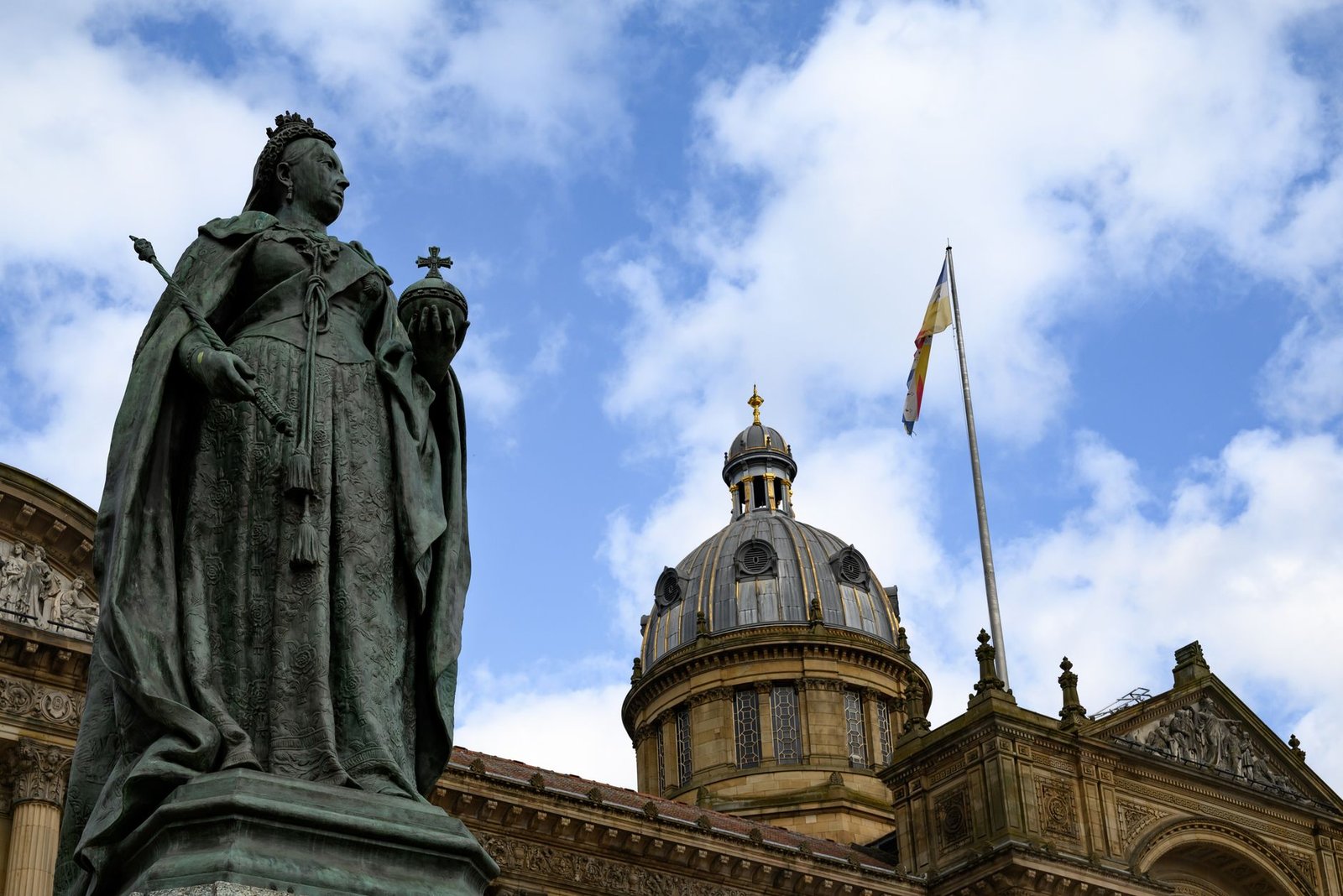 A historic statue with ornate architecture and a flag in Birmingham, England under a partly cloudy sky.