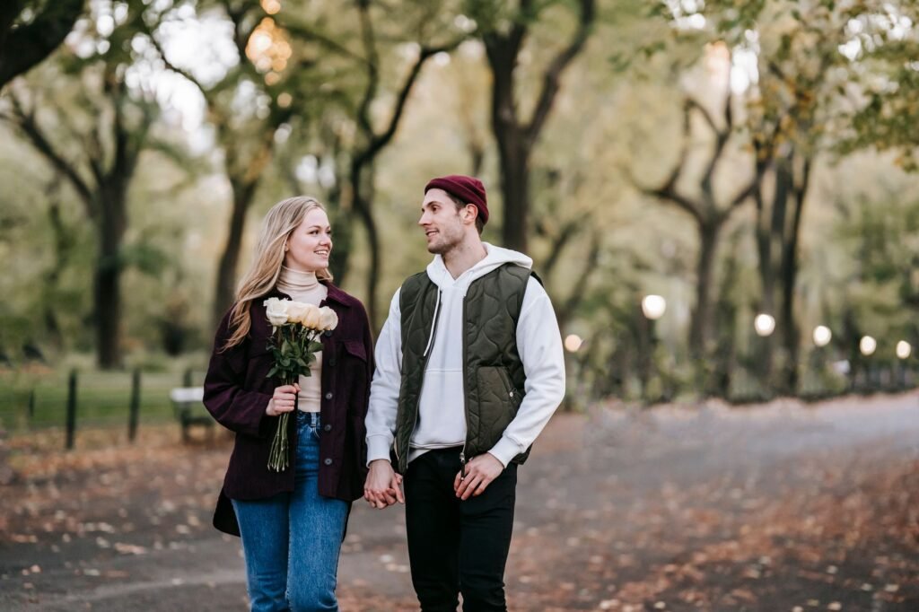 Cheerful couple with bouquet of fresh white roses promenading in park with green trees and lights