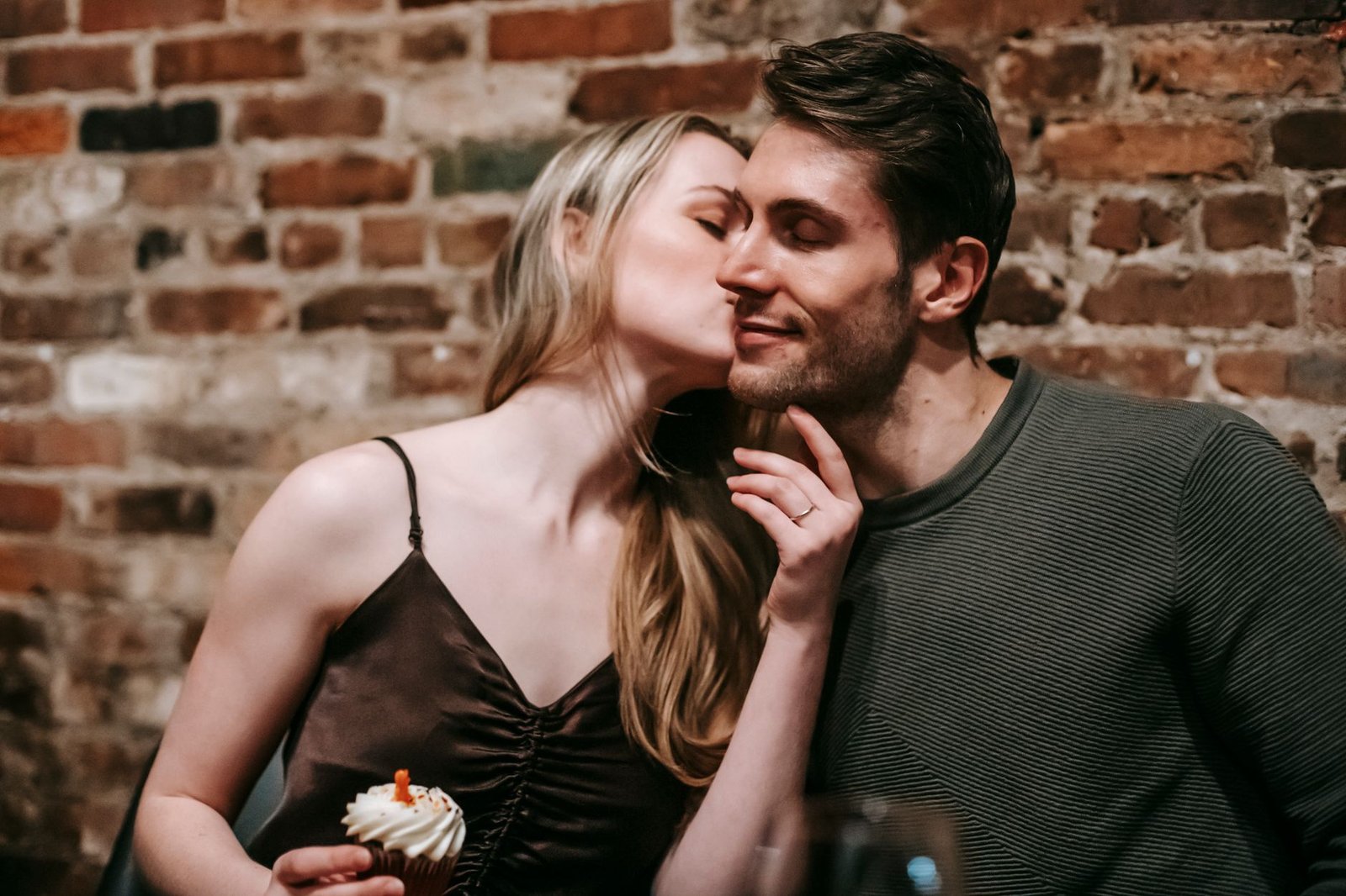 Young couple in elegant clothes kissing in restaurant on date while celebrating birthday with cupcake and candle near brick w
