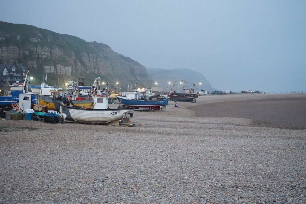 A serene scene of fishing boats at dusk on a pebble beach with cliffs in the background.