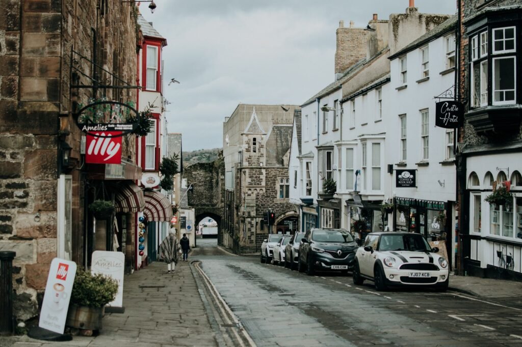 A quaint British street lined with shops and parked cars leading to an old stone gate.