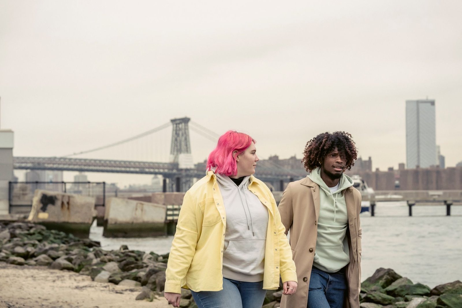 Multiethnic couple in outerwear strolling together on shore with stones near calm river against bridge and residential buildi