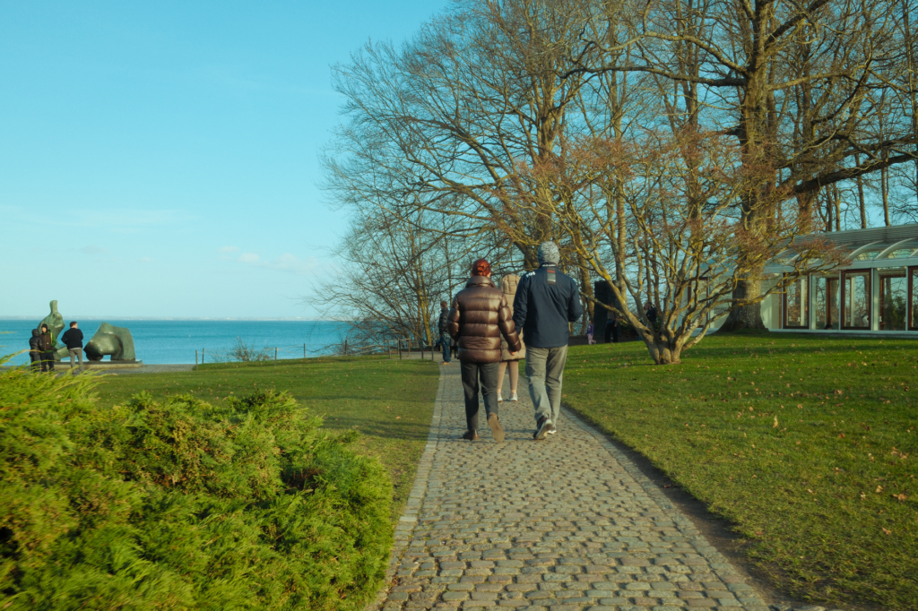 A couple walks on a park path by the seaside, enjoying a bright day.
