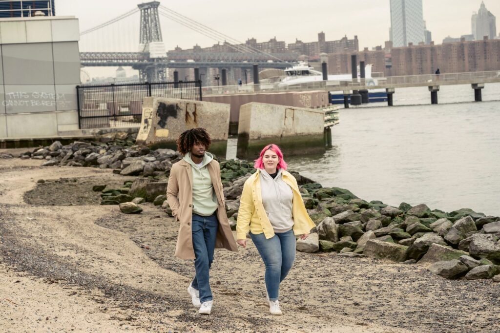 Full body of multiracial couple in jeans strolling on embankment with stones against modern bridge and buildings located in c