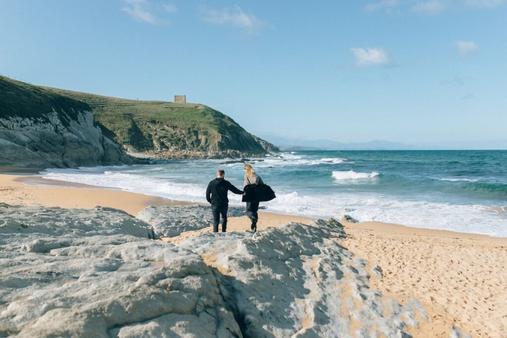 A couple walks hand in hand along a picturesque beach with rocky cliffs and waves, under a clear blue sky.