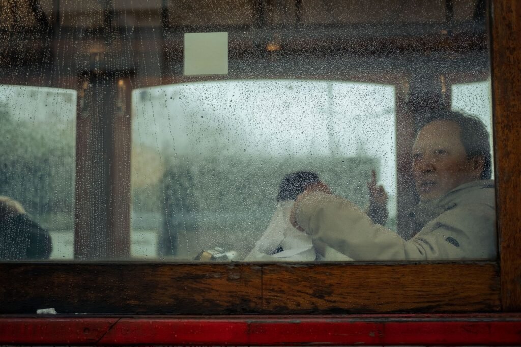 A man wearing a beige jacket looking out through a wet window on a rainy day.