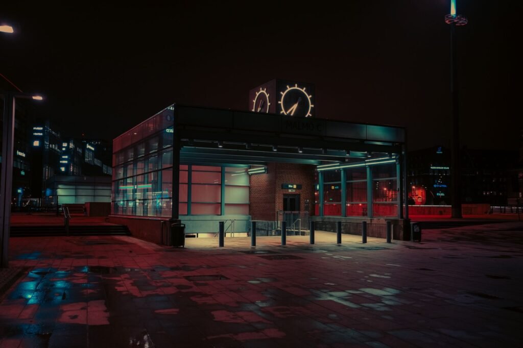 A moody, futuristic view of Malmö metro station at night with neon lights and rain reflections.