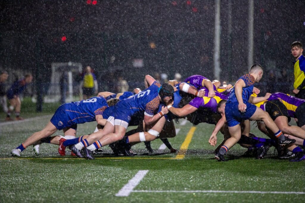 Dramatic rugby scrum at night in Manchester rain, showcasing teamwork and athleticism.