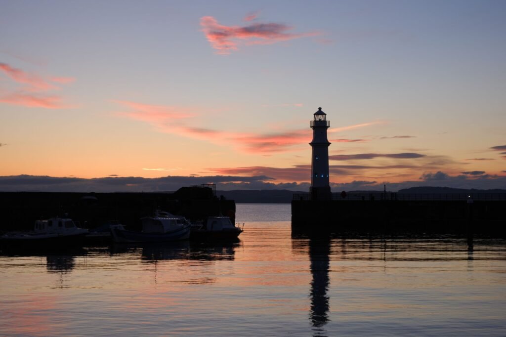 Serene sunset view of the Edinburgh Harbour featuring a lighthouse and boats reflecting in the water.