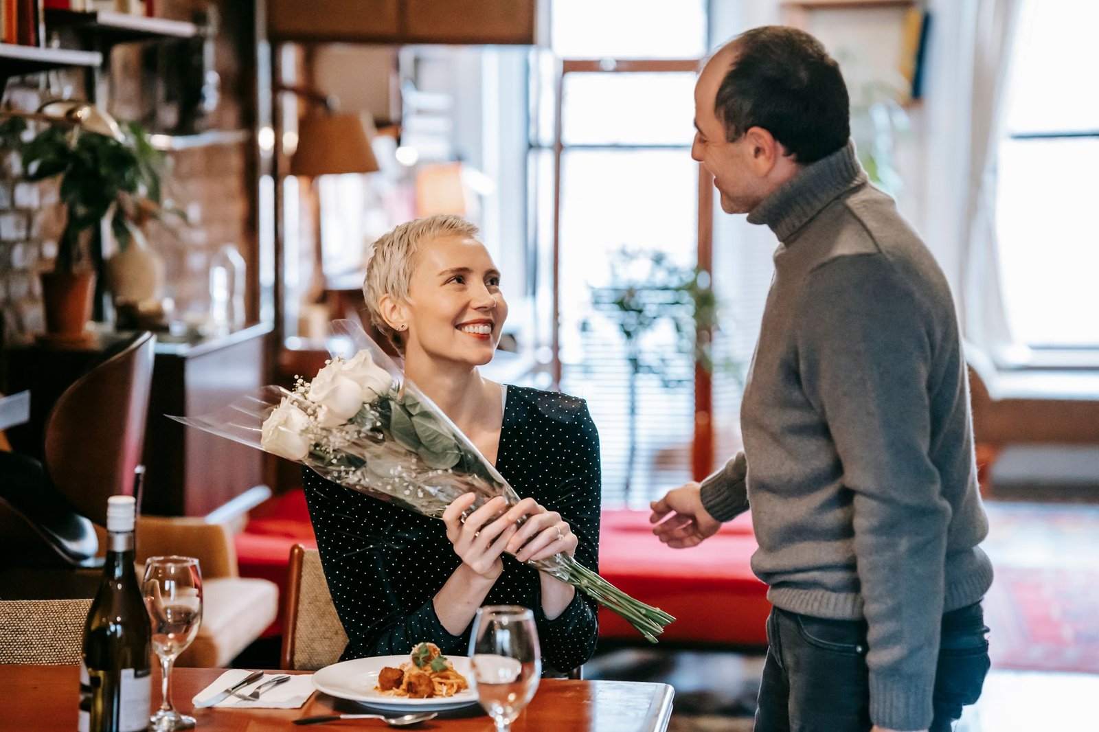 Happy couple having romantic dinner while man giving bouquet of white roses to woman near table with wine bottle and glasses