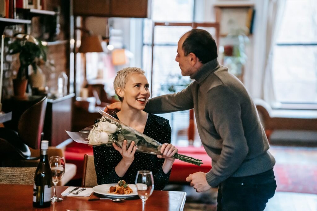 Couple having romantic dinner while man giving bouquet of white roses to woman near table with plate with pasta near wine bot