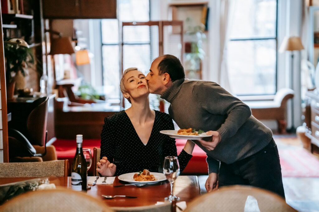 Gentle husband with plate of meal kissing wife having romantic dinner with bottle of wine at home