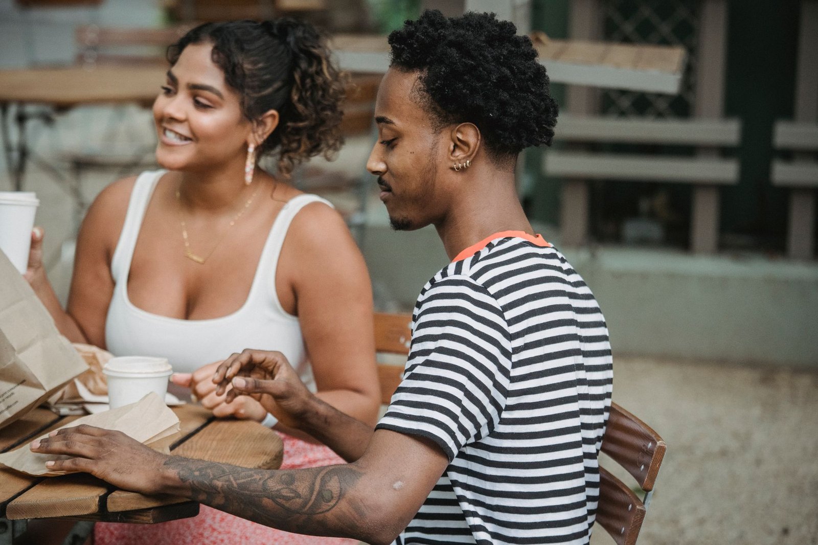 Happy young Hispanic woman and African American man in casual clothes resting on cafe terrace and drinking takeaway coffee du