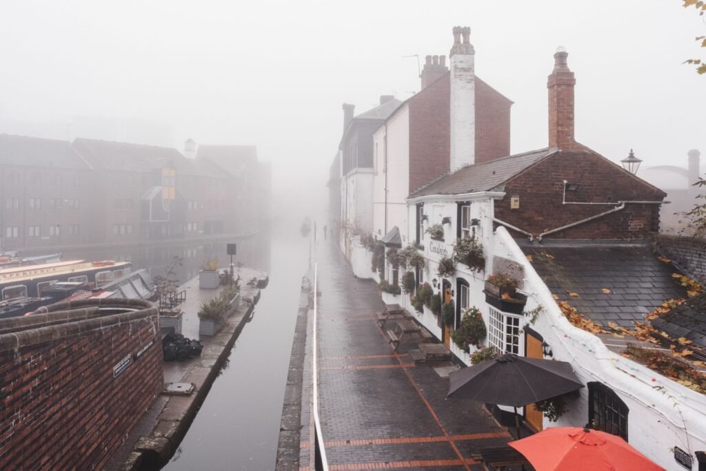 Narrow water channel flowing along old fashioned buildings and terraces of cafes with tables and parasols in city on foggy we