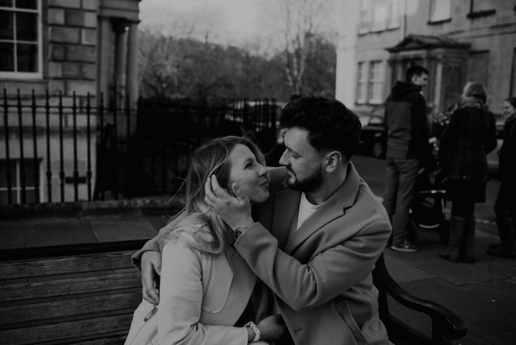 A romantic couple shares a tender moment on a bench in Bath, UK city street.