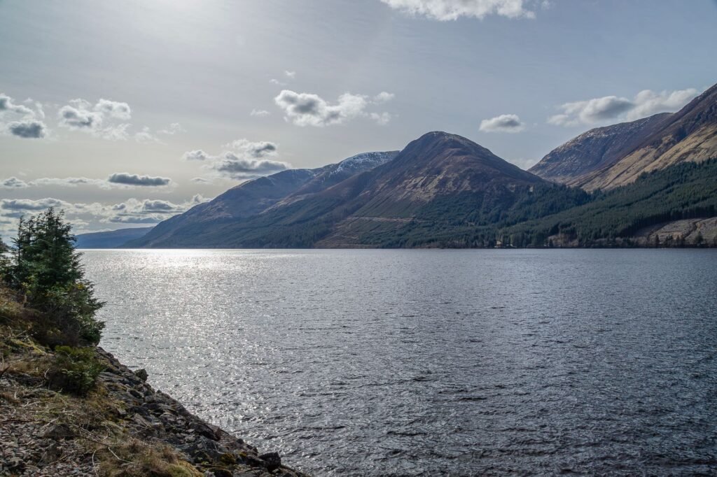 Scenic view of Loch Ness with surrounding mountains in Scotland under a cloudy sky.