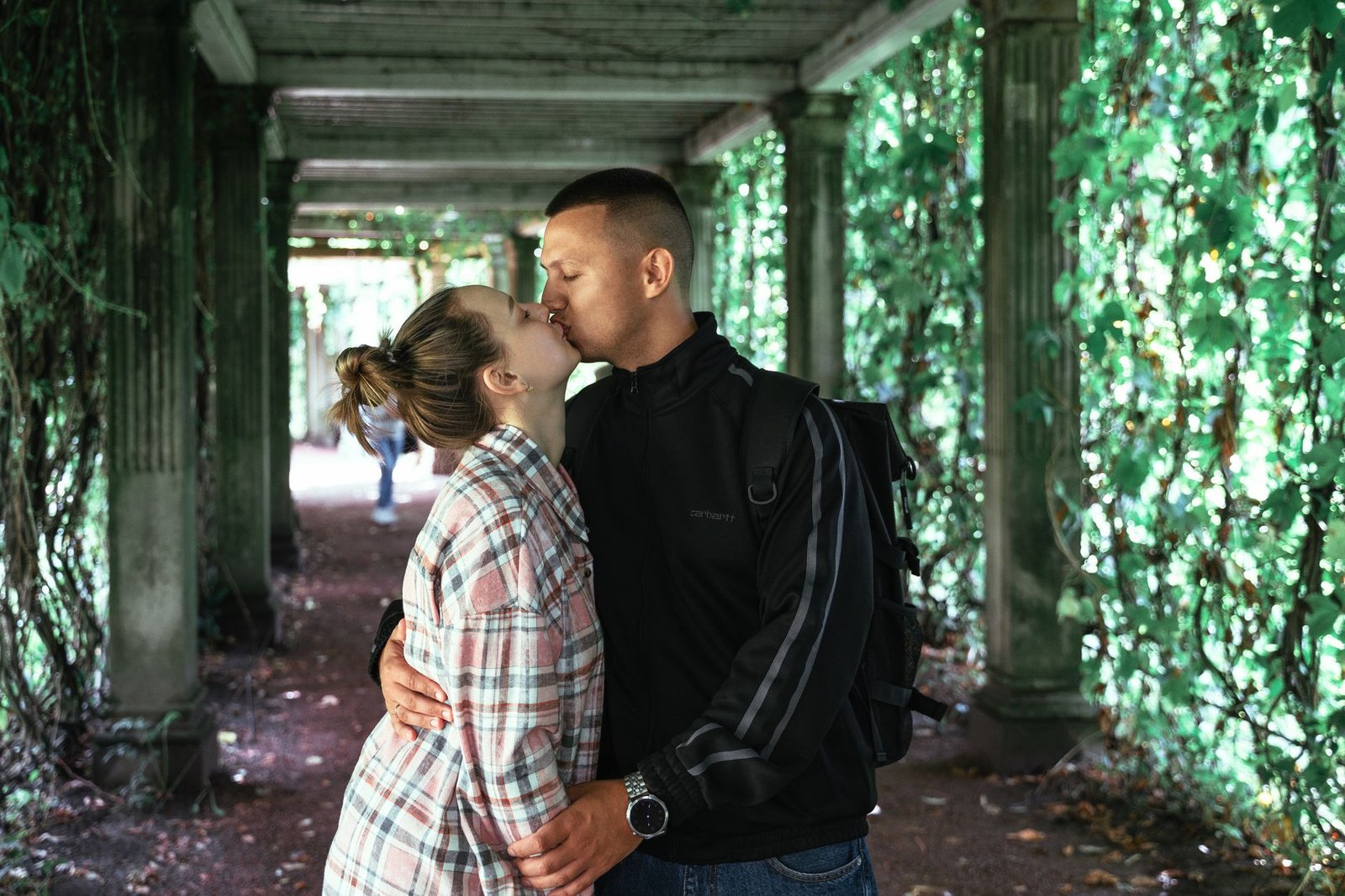 A couple shares a romantic kiss in a lush, vine-covered corridor on a bright day.