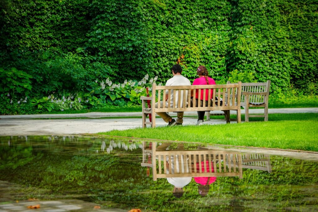 A couple sitting on a wooden bench, enjoying a serene green park with lush greenery and a reflecting pond.