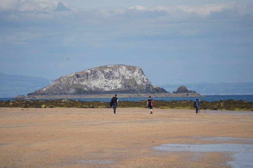 A serene beach scene with people walking towards Bass Rock in North Berwick, Scotland.