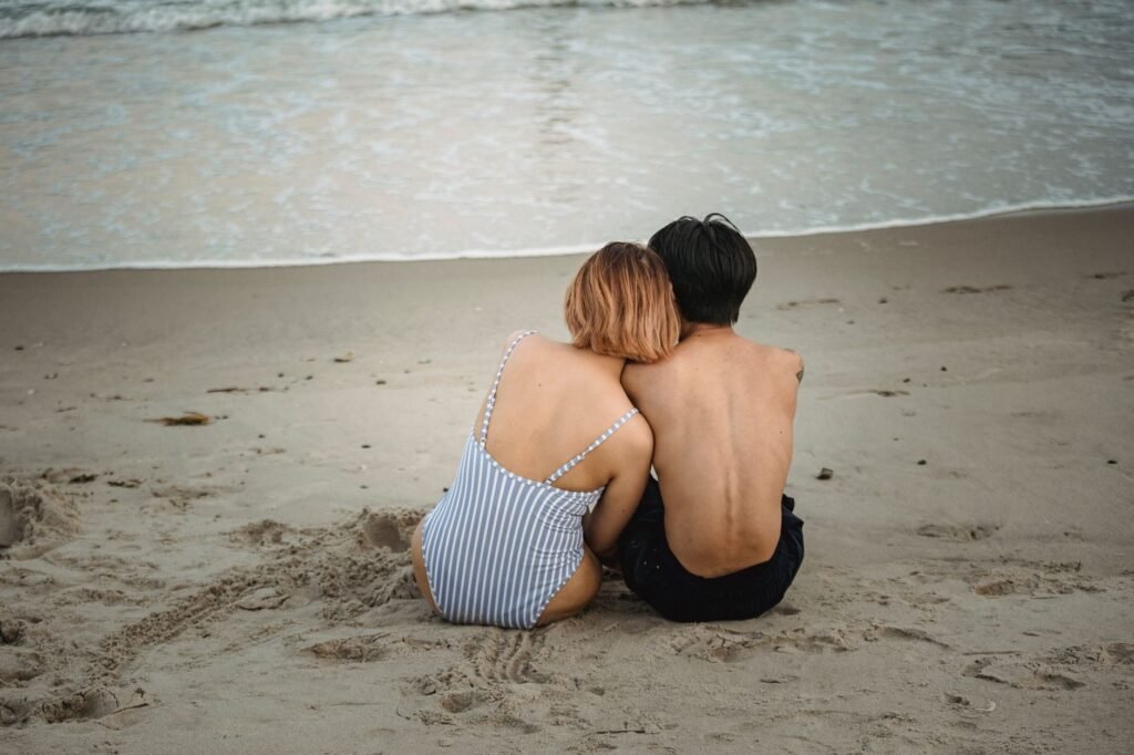 A couple enjoys a romantic moment sitting close on a sandy beach at sunset, overlooking the sea.